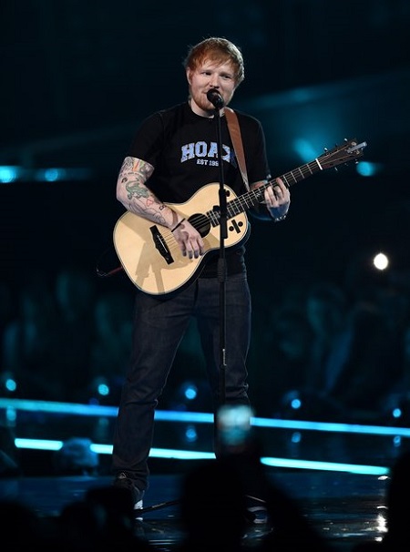 Ed Sheeran on stage in front of the mic with a guitar in his hands.